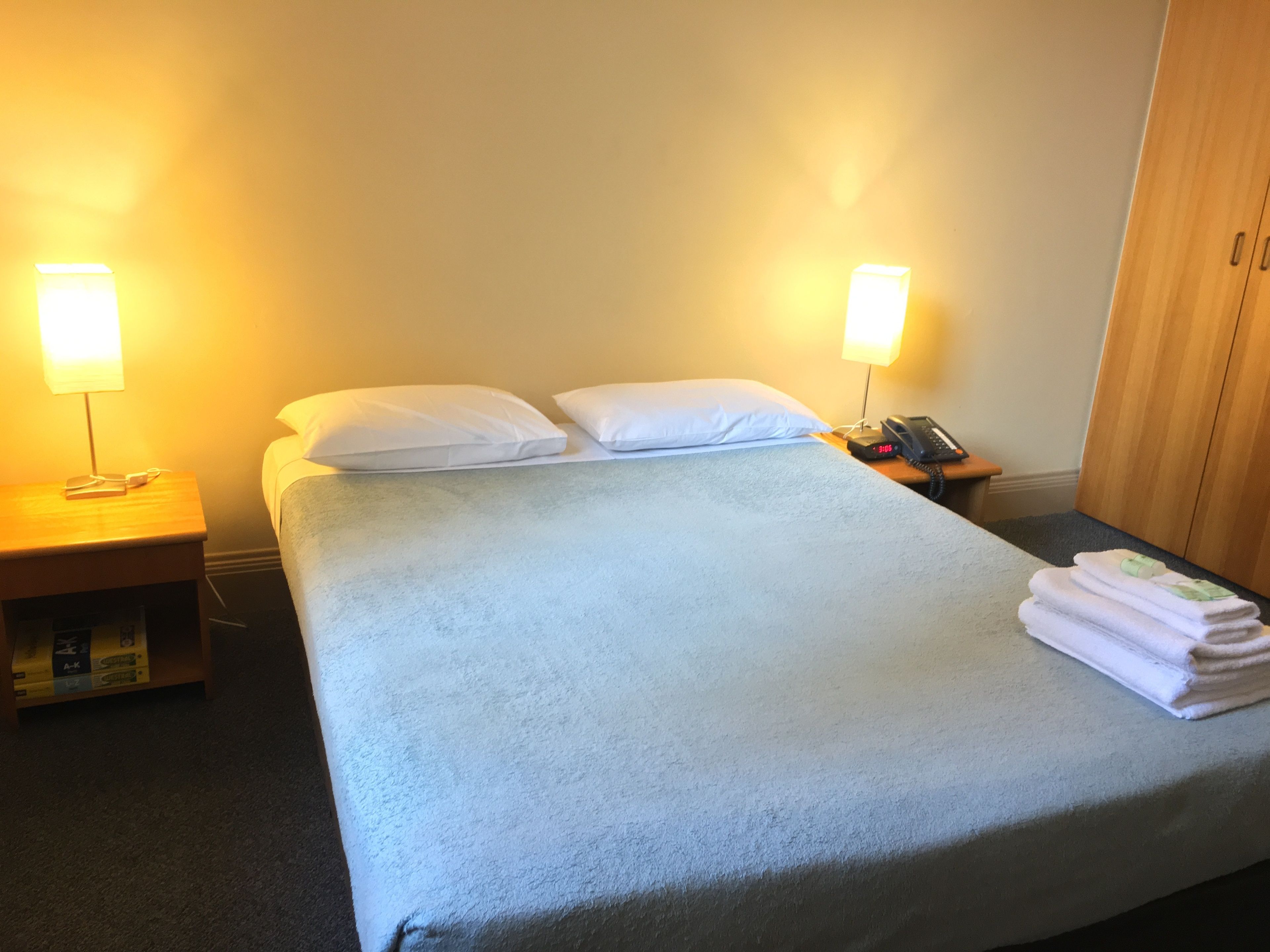 Habitación de hotel con cama, dos lámparas de noche, mesita de noche con teléfono y libros, y una pila de toallas sobre la cama.