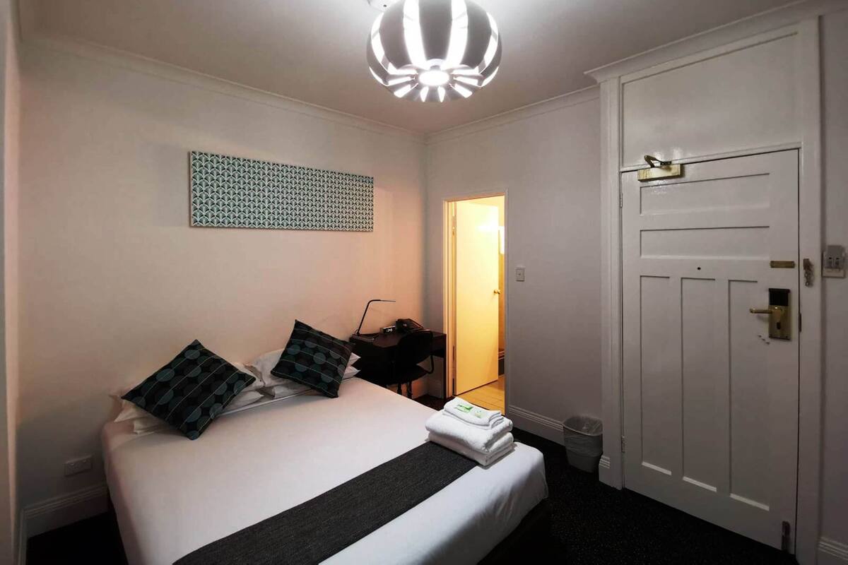 A hotel room with a bed, a desk, a chair, a white door, and a ceiling light fixture.