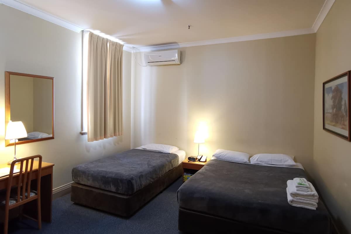A hotel room with two beds, a desk, a chair, a lamp, an air conditioning unit, and a painting on the wall.