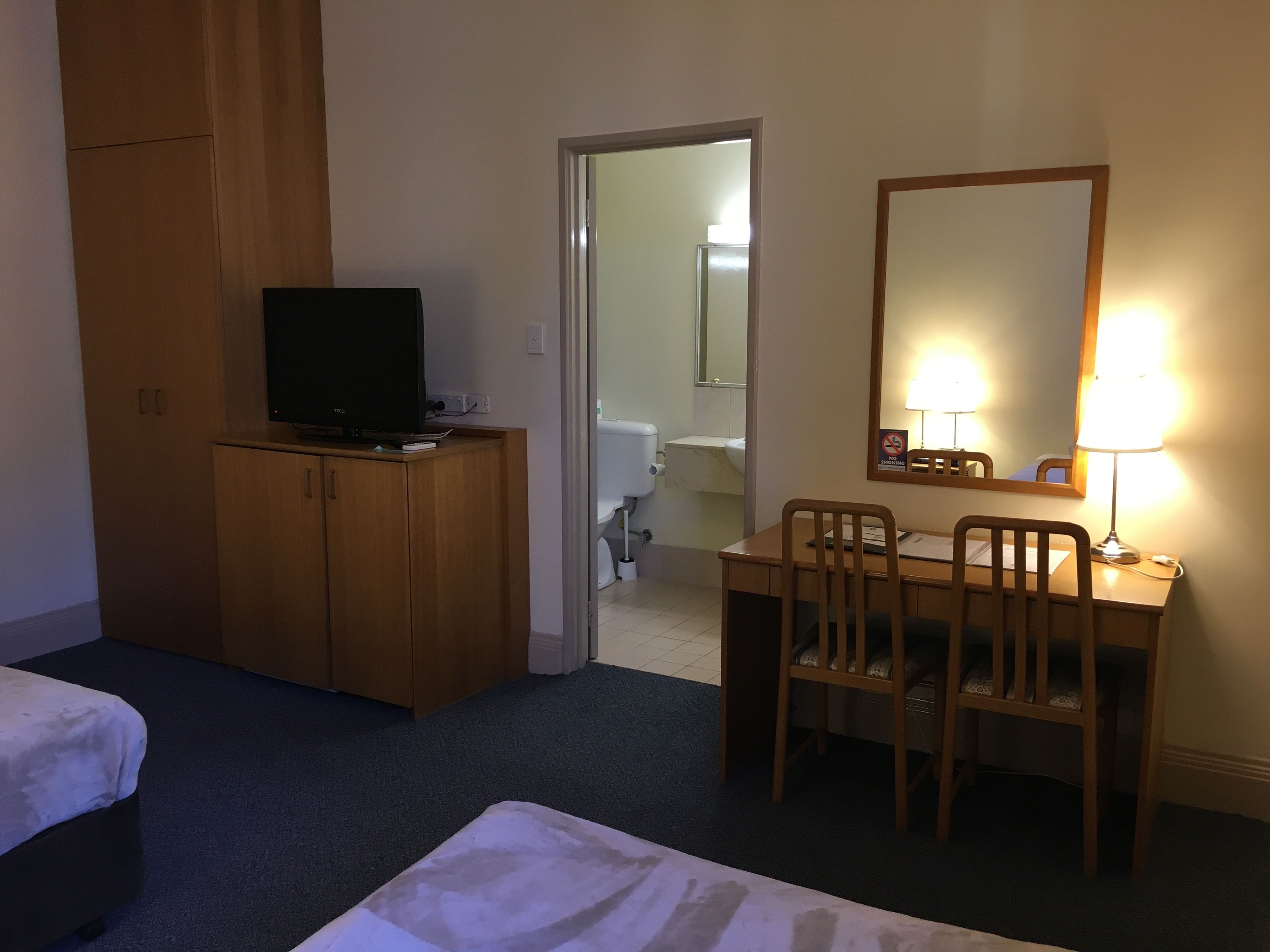 Habitación de hotel con televisión sobre un mueble de madera, un escritorio con silla, un espejo y una mesita de noche.