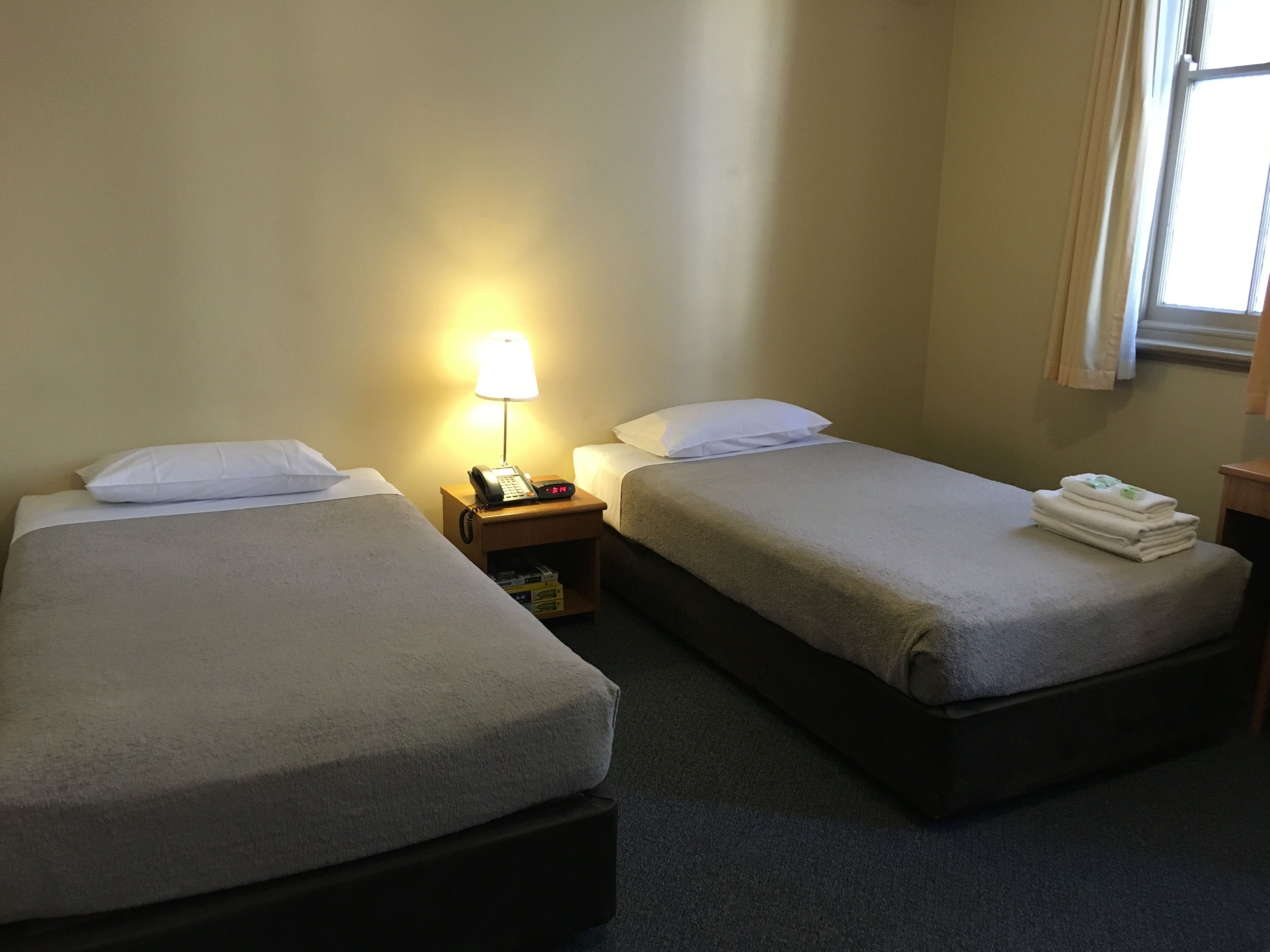 A hotel room with two single beds, a bedside table with a lamp, and a small shelf with a telephone.