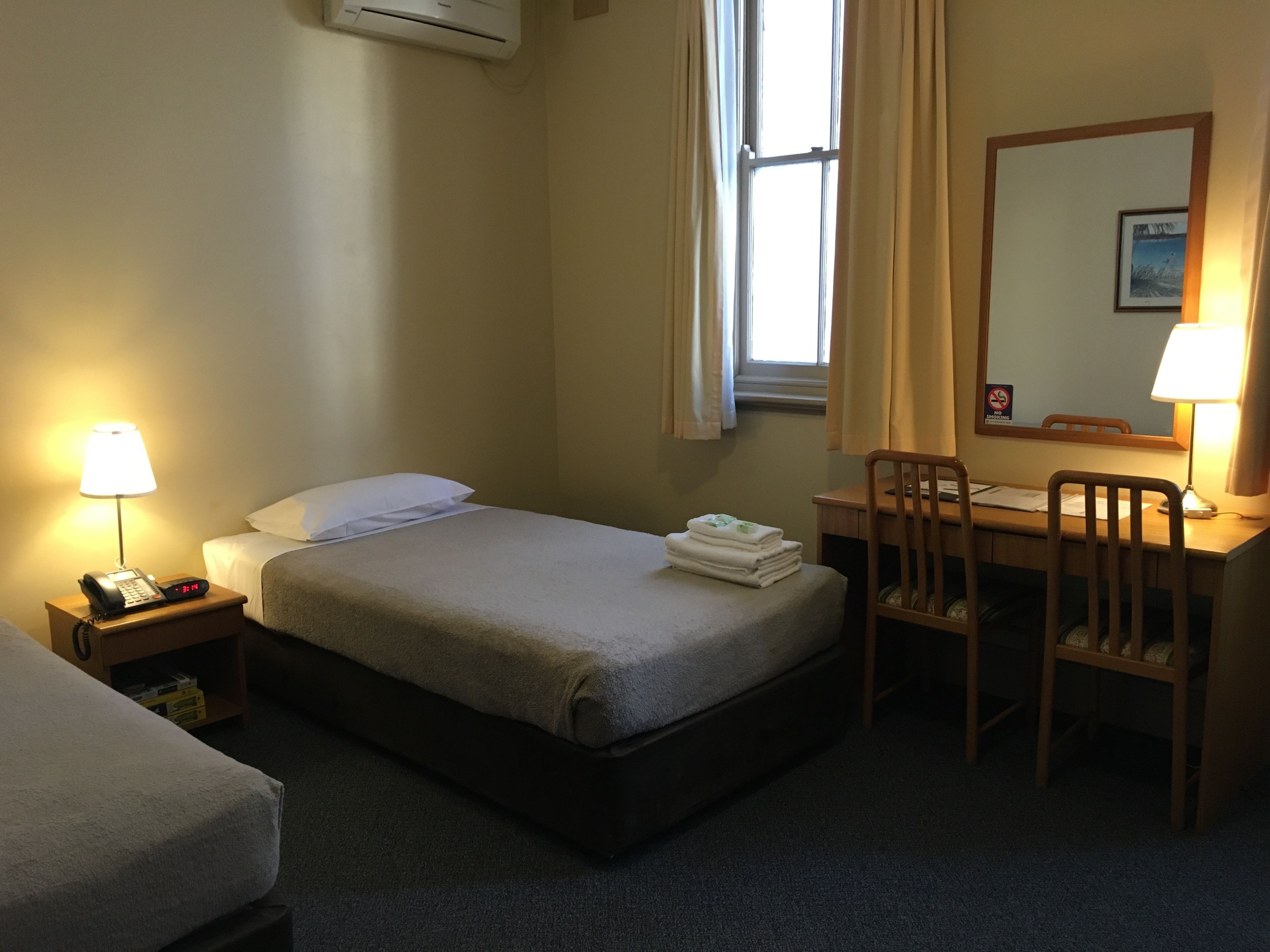 Habitación de hotel con cama, escritorio y silla, mesita de noche con lámpara, y ventana con cortinas.