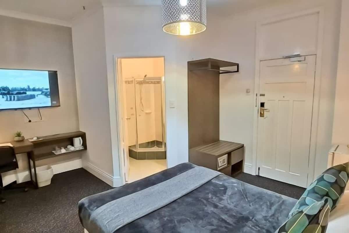 A hotel room with a bed, a desk, a TV, a shower, and a door.