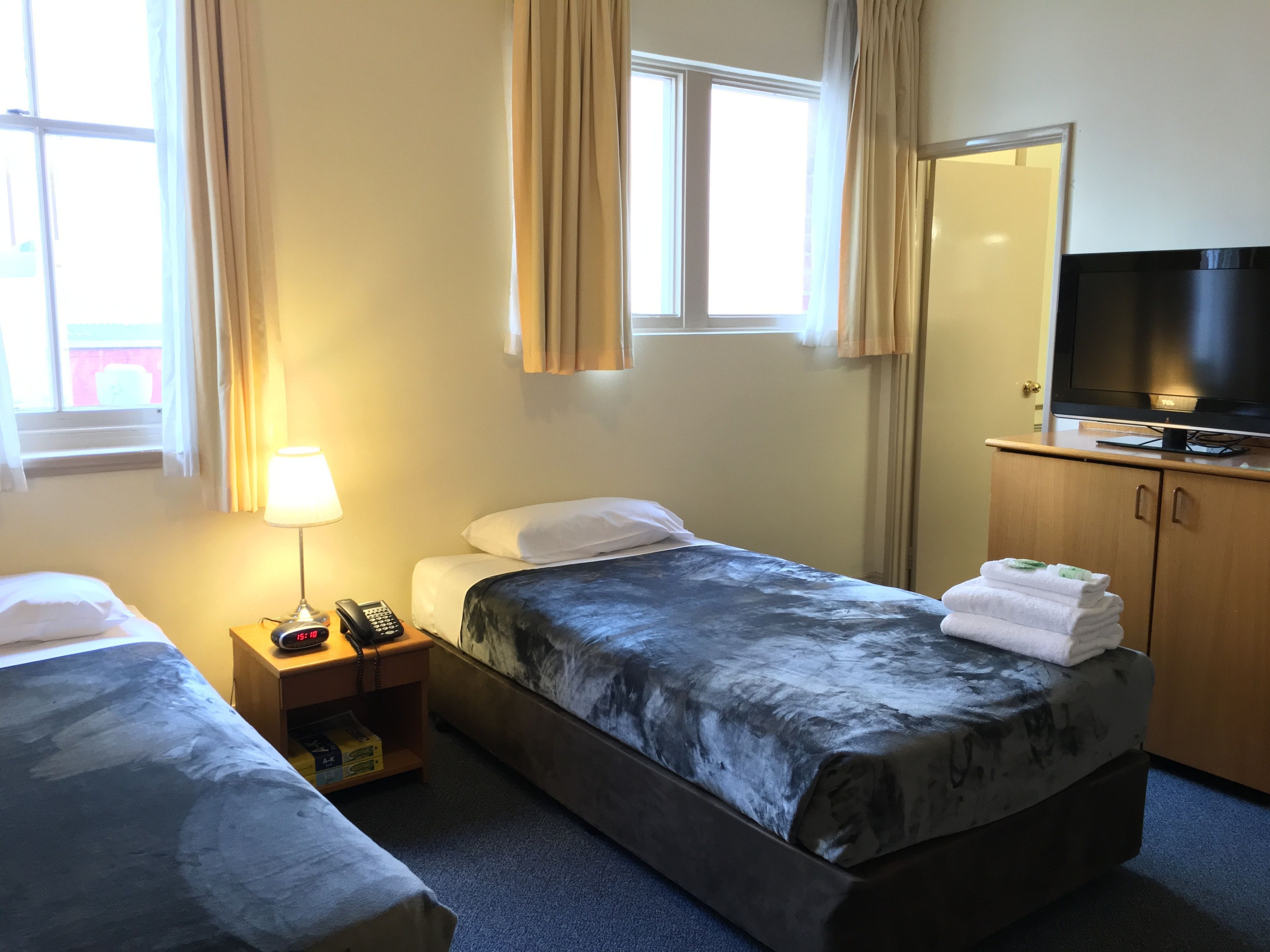 Habitación de hotel con dos camas individuales, un televisor sobre un mueble de madera, una mesita de noche con lámpara y teléfono, y una ventana con cortinas.