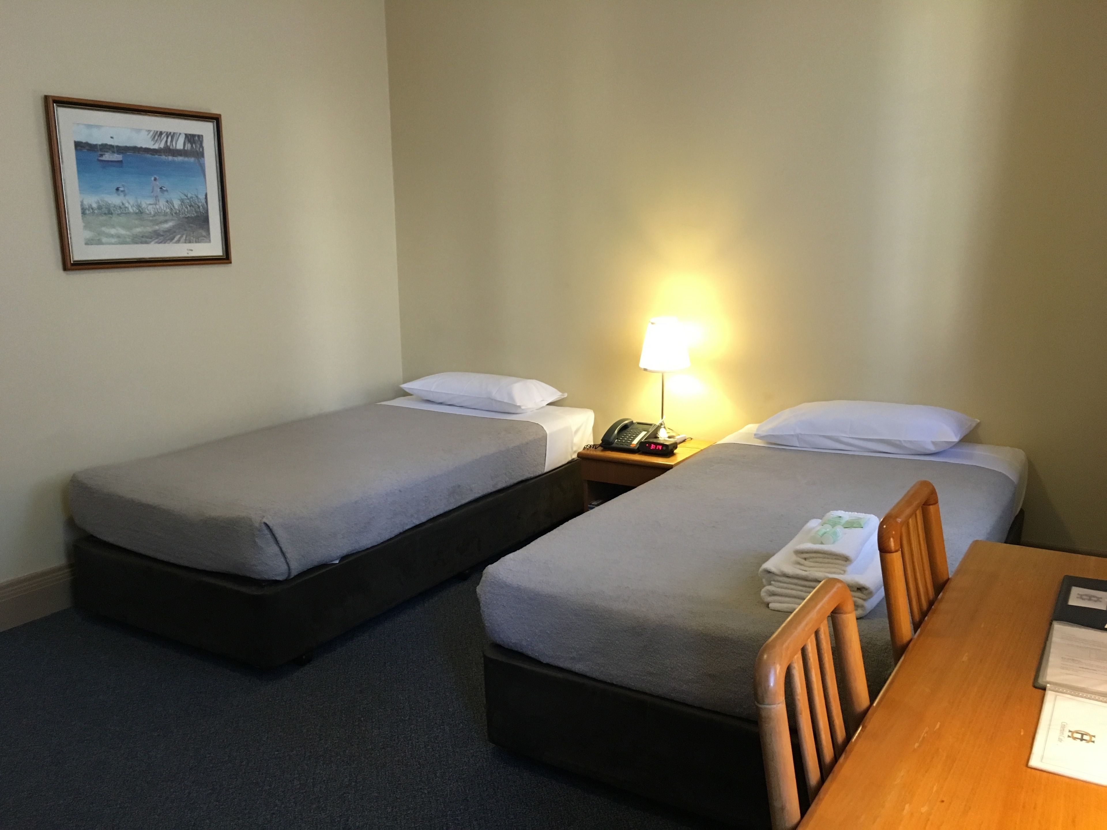 Quarto de hotel com duas camas de solteiro, uma mesa de madeira, uma mesa de cabeceira com luminária e um quadro na parede.