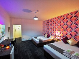 Camera d'albergo con due letti, una scrivania, una sedia, una ventola a soffitto e un muro decorato.