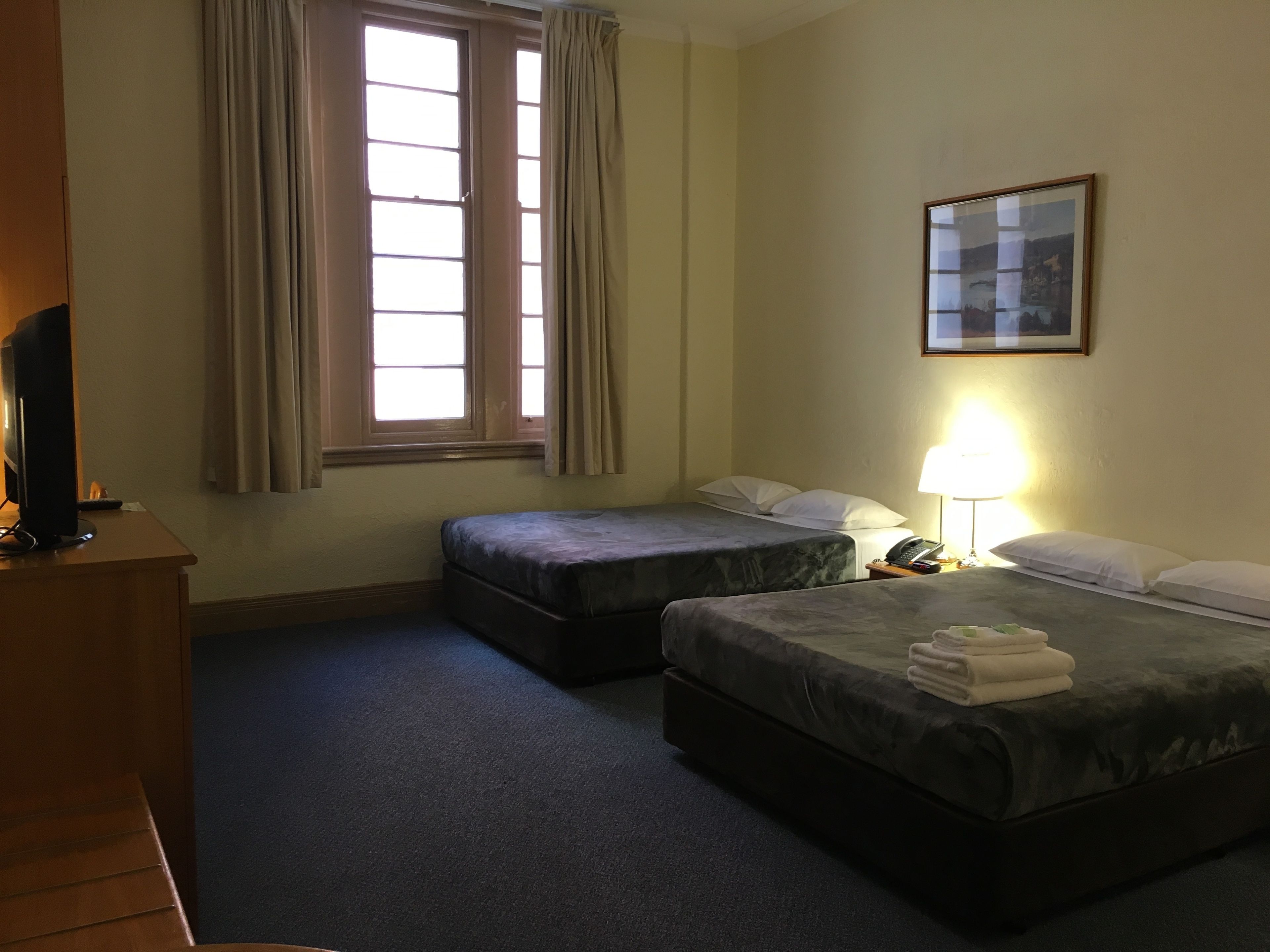 A hotel room with two beds, a desk with a computer, a window with curtains, a lamp, and a framed picture on the wall.