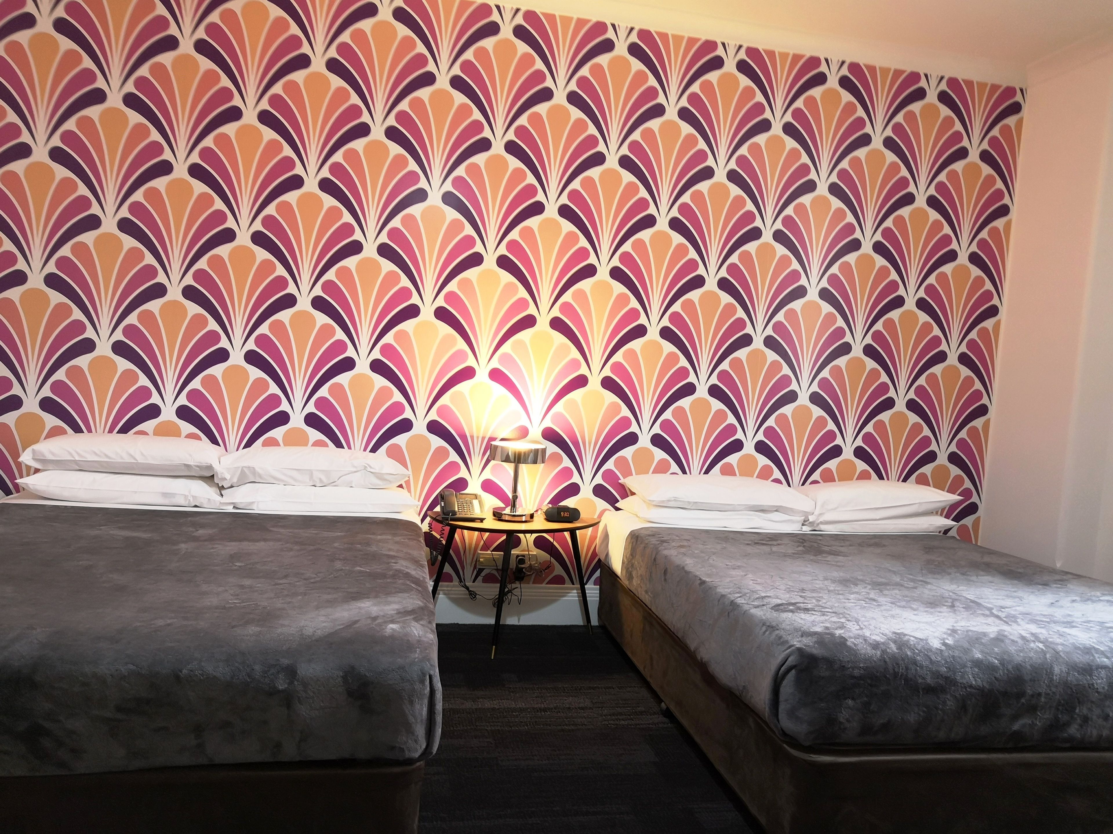 Quarto com duas camas, uma mesa pequena com abajur e parede estampada.