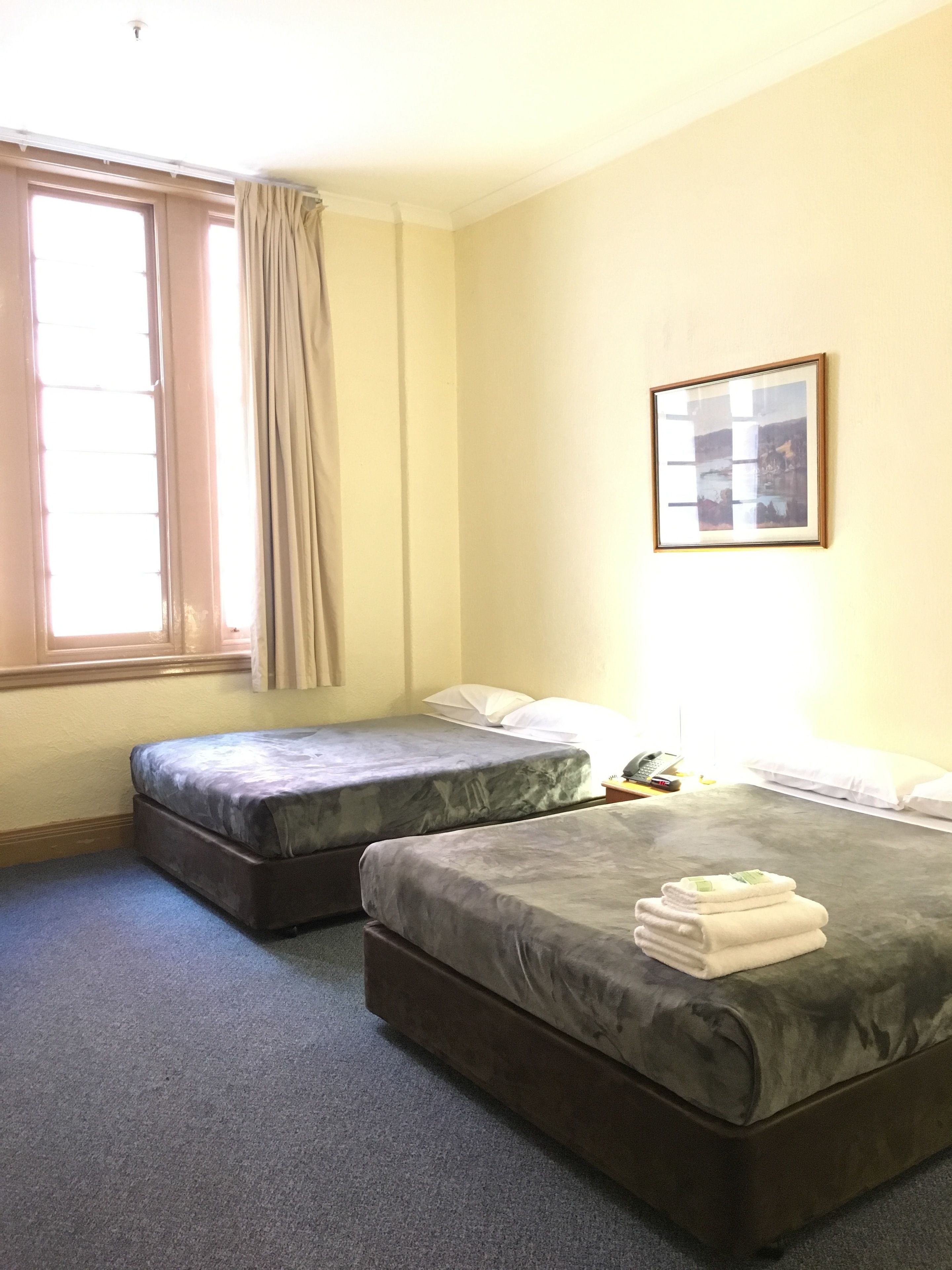A hotel room with two beds, a window with curtains, a framed picture on the wall, and a small table with a phone and a book.