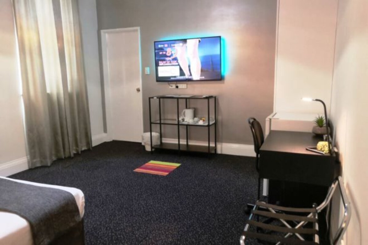 A hotel room with a TV mounted on the wall, a small table with chairs, a shelf with items, and a door.