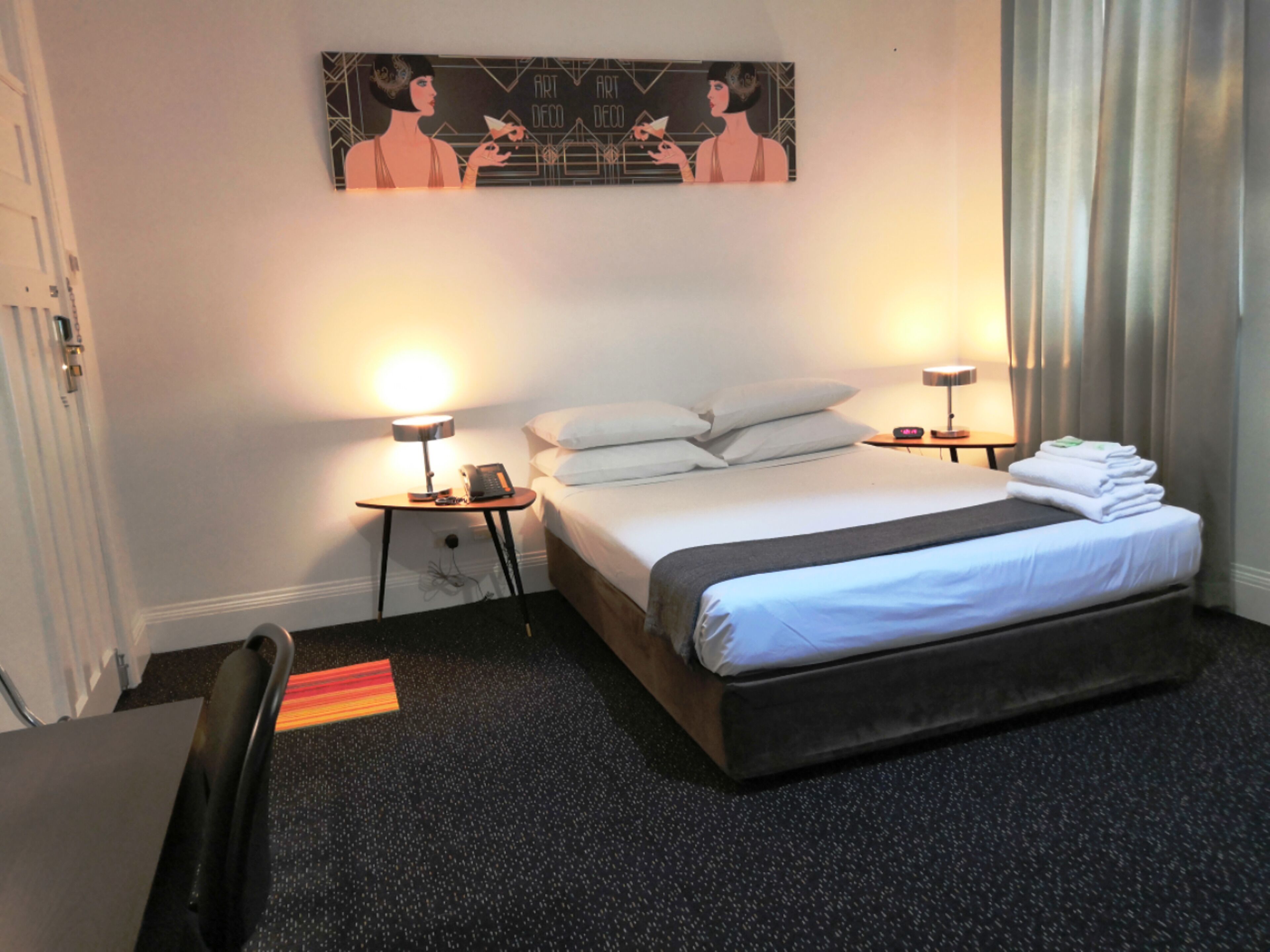 A hotel room with a bed, two bedside tables with lamps, a painting on the wall, and a chair.