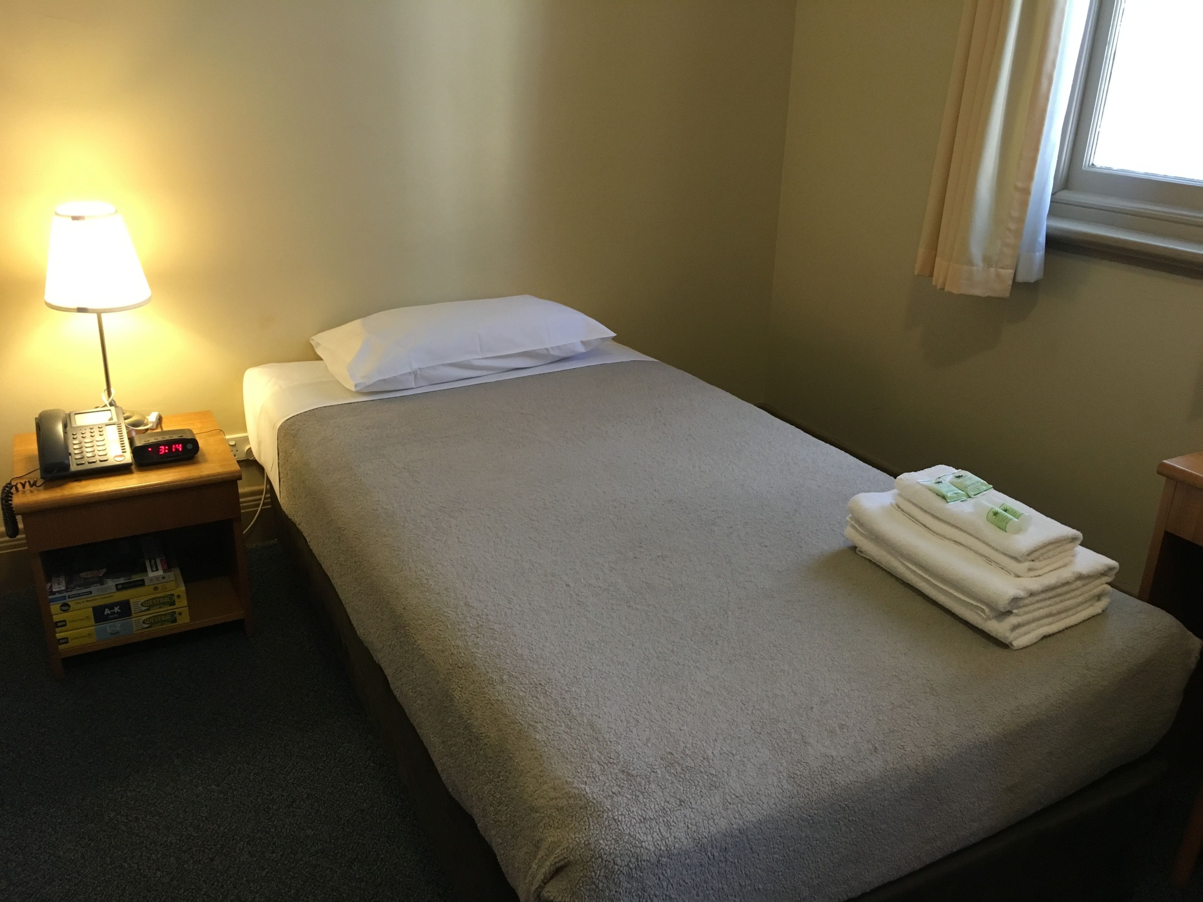 A hotel room with a bed, a nightstand with a lamp, a phone, and a small shelf with toiletries.