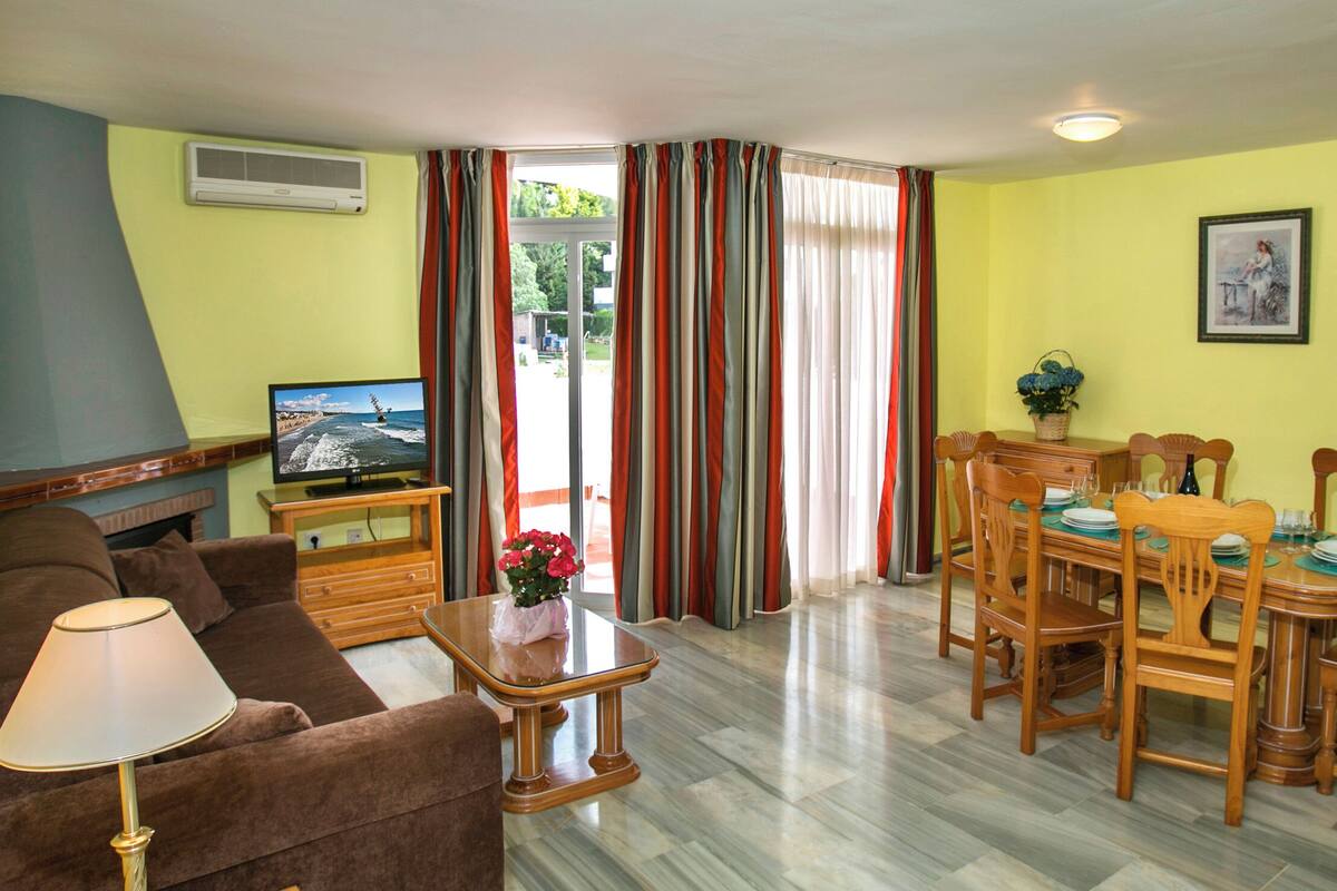 A living room with a brown sofa, a wooden coffee table with a vase of flowers, a TV on a wooden stand, and a dining area with a wooden table and chairs.