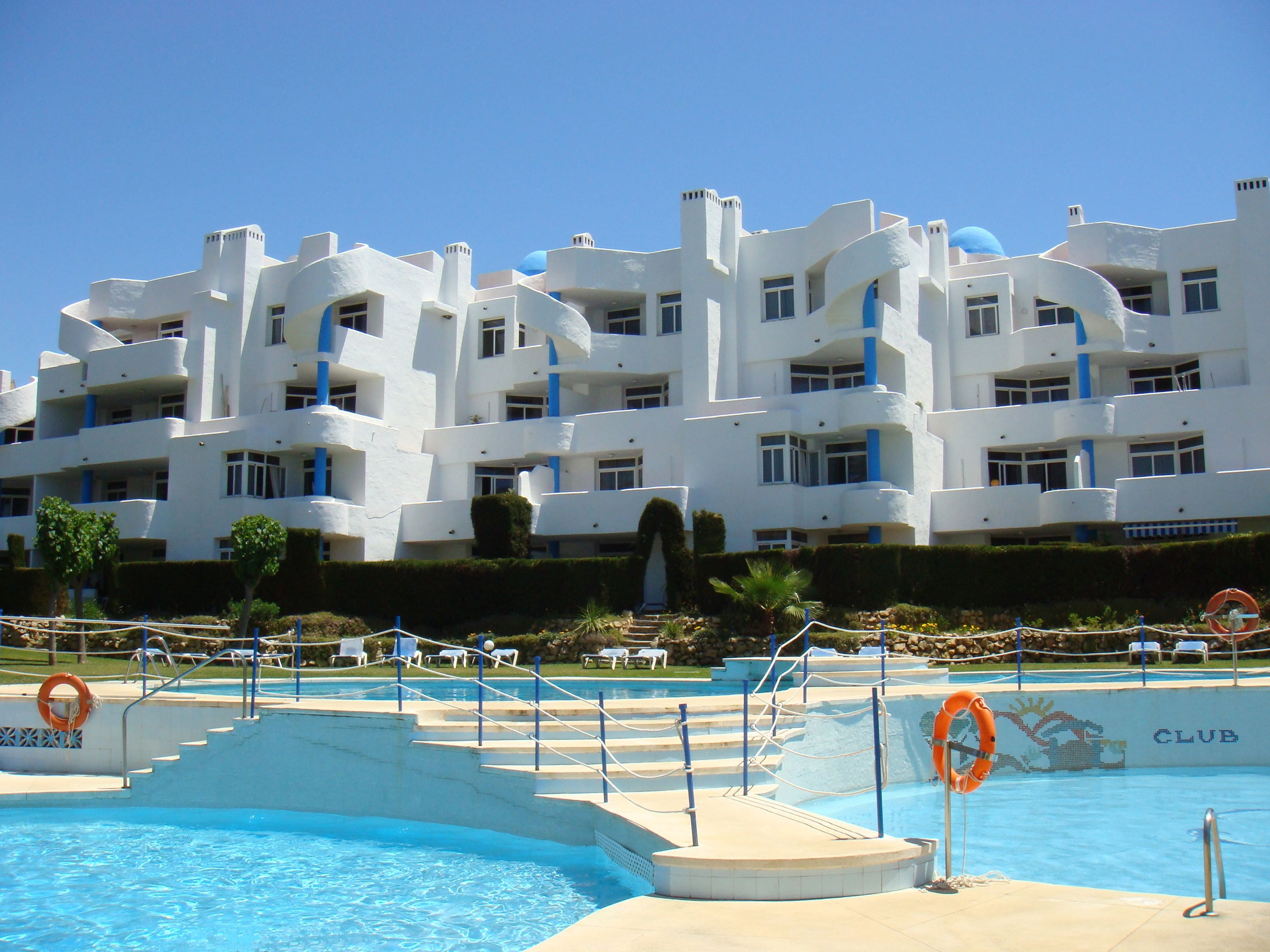 A hotel with a pool area, featuring a clear blue pool, steps leading into the pool, and a lifebuoy near the pool's edge.
