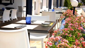 An outdoor dining area with white chairs and tables, blue glassware, and a row of pink flowers. - Apartamentos Ona Campanario (Mijas)