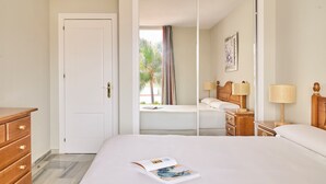 A hotel room with a bed, a wooden dresser, a nightstand with lamps, and a window with a view of palm trees. - Apartamentos Ona Campanario (Mijas)