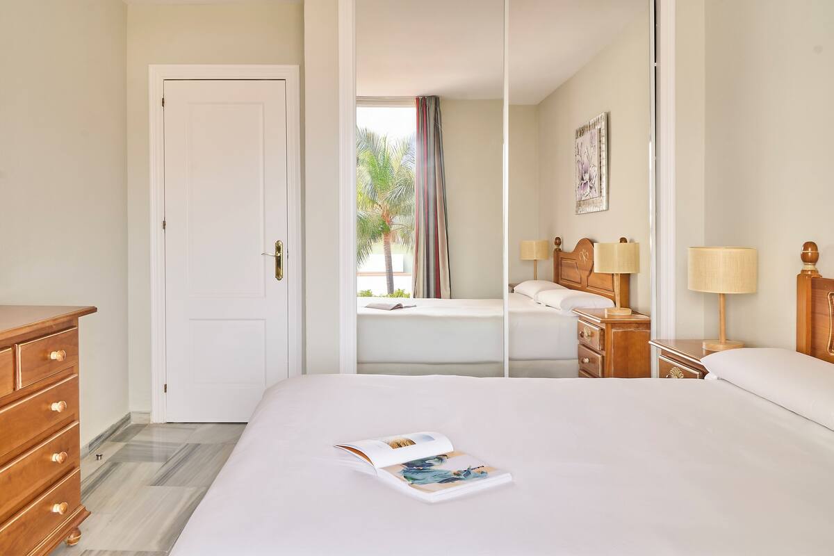 A hotel room with a bed, a wooden dresser, a nightstand with lamps, and a window with a view of palm trees.