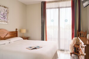 Una camera d'albergo con un letto grande, un comodino in legno con una lampada, una cassettiera in legno e una finestra con tende.