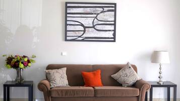 Una sala de estar con un sofá marrón, dos mesitas auxiliares, un jarrón de flores y un cuadro abstracto colgado en la pared.