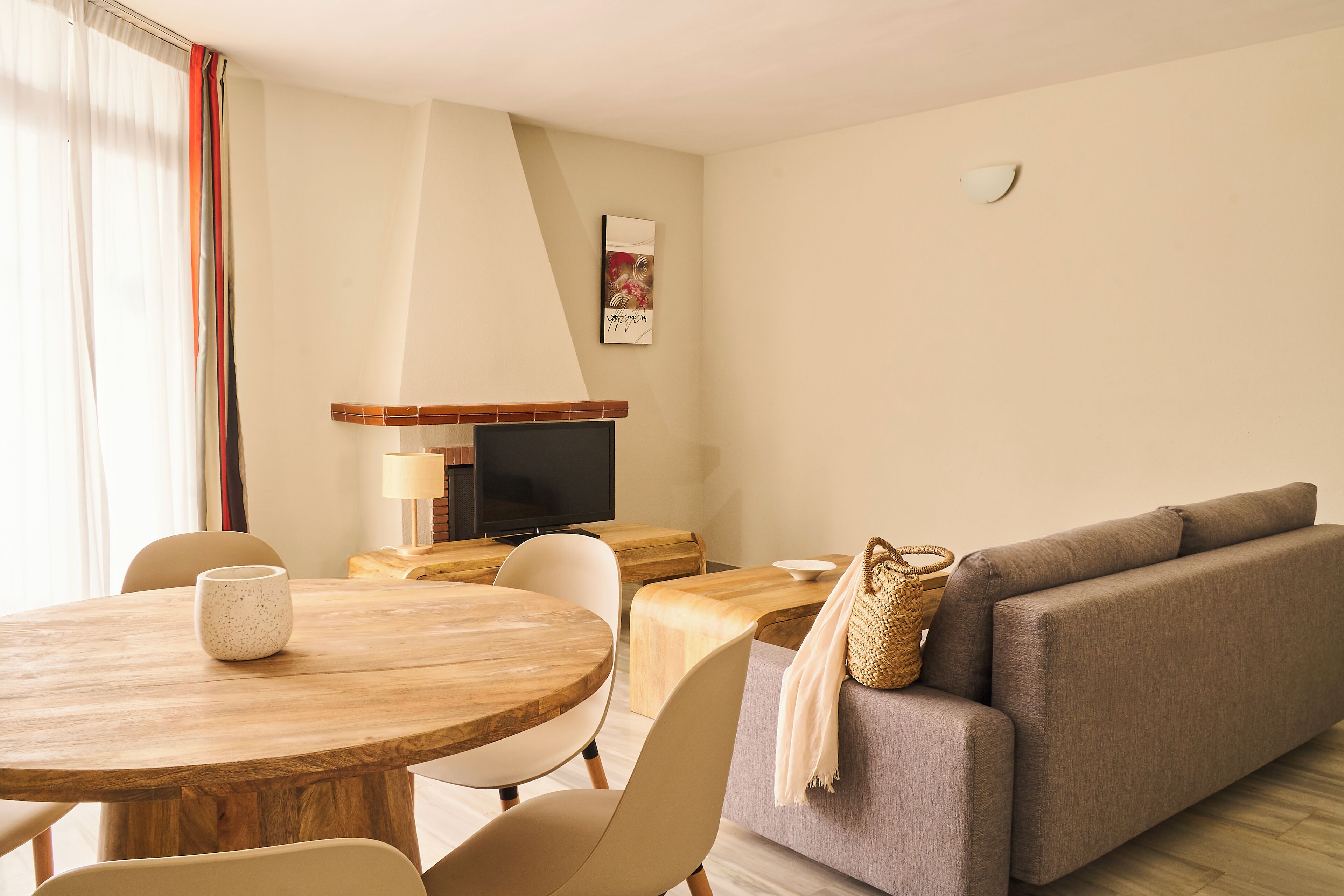 Una sala de estar moderna con una mesa redonda de madera, sillas color beige, un sofá gris y un televisor sobre un mueble de madera.