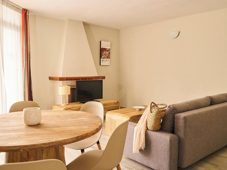 Una sala de estar moderna con una mesa redonda de madera, sillas color beige, un sofá gris y un televisor sobre un mueble de madera.