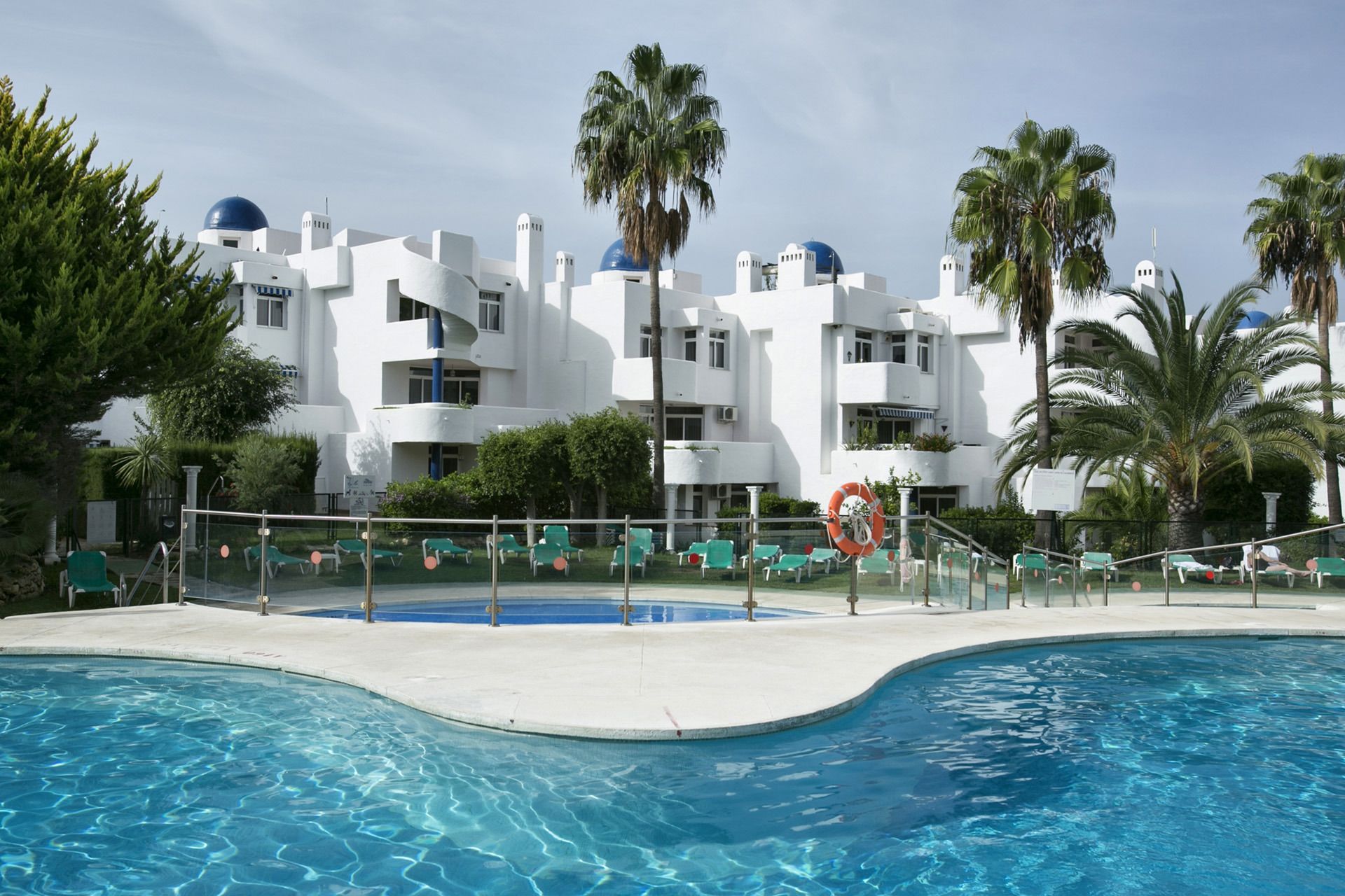 Un hôtel moderne doté d’un espace piscine, avec des bâtiments blancs surmontés de coupoles bleues, des palmiers et des chaises longues.