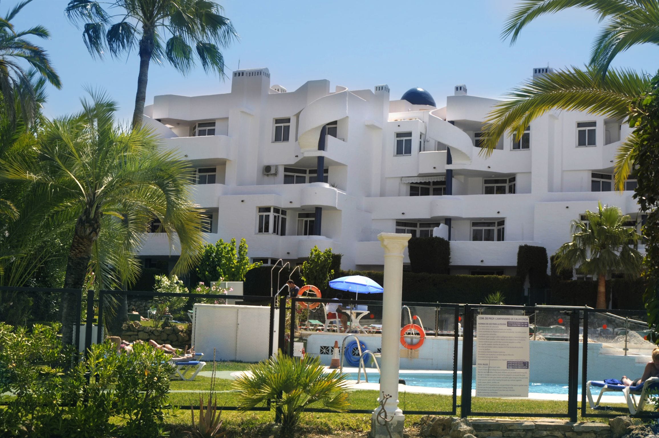 A modern hotel with a pool area, surrounded by palm trees and a fenced poolside.