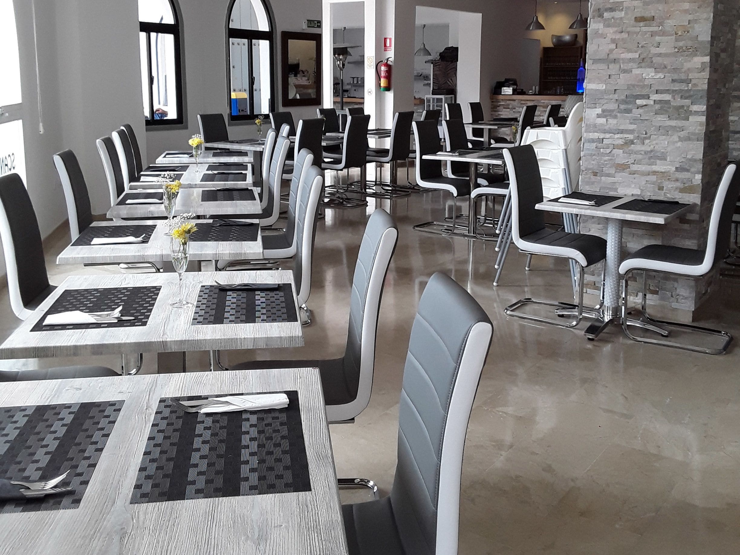 Un restaurant moderne doté de nombreuses tables et chaises, d’un mur décoratif en pierre et d’une cuisine ouverte visible au fond de la salle.