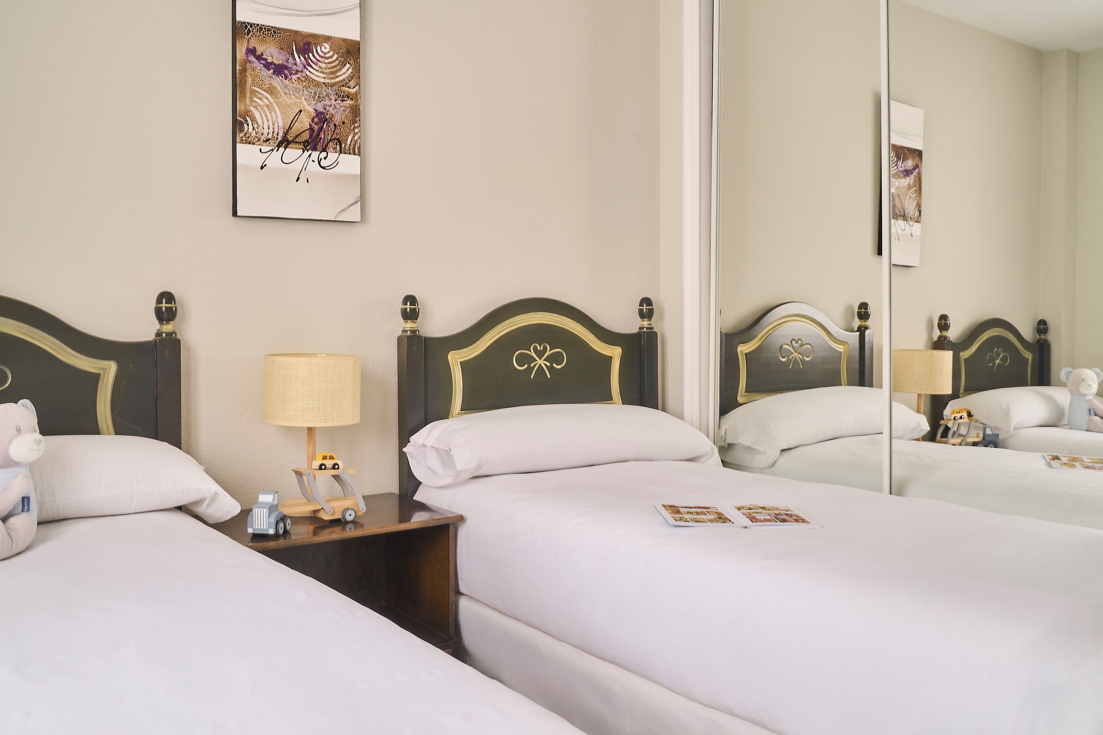 A hotel room with two beds, a nightstand with a lamp, a teddy bear, and a mirror reflecting the room.