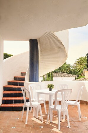 A terrace with white plastic chairs and a table, a spiral staircase, and a view of palm trees and buildings. - Apartamentos Ona Campanario (Mijas)