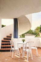 A terrace with white plastic chairs and a table, a spiral staircase, and a view of palm trees and buildings.