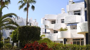 A modern hotel with white exterior walls, balconies, and lush greenery including palm trees and red-leafed plants. - Apartamentos Ona Campanario (Mijas)