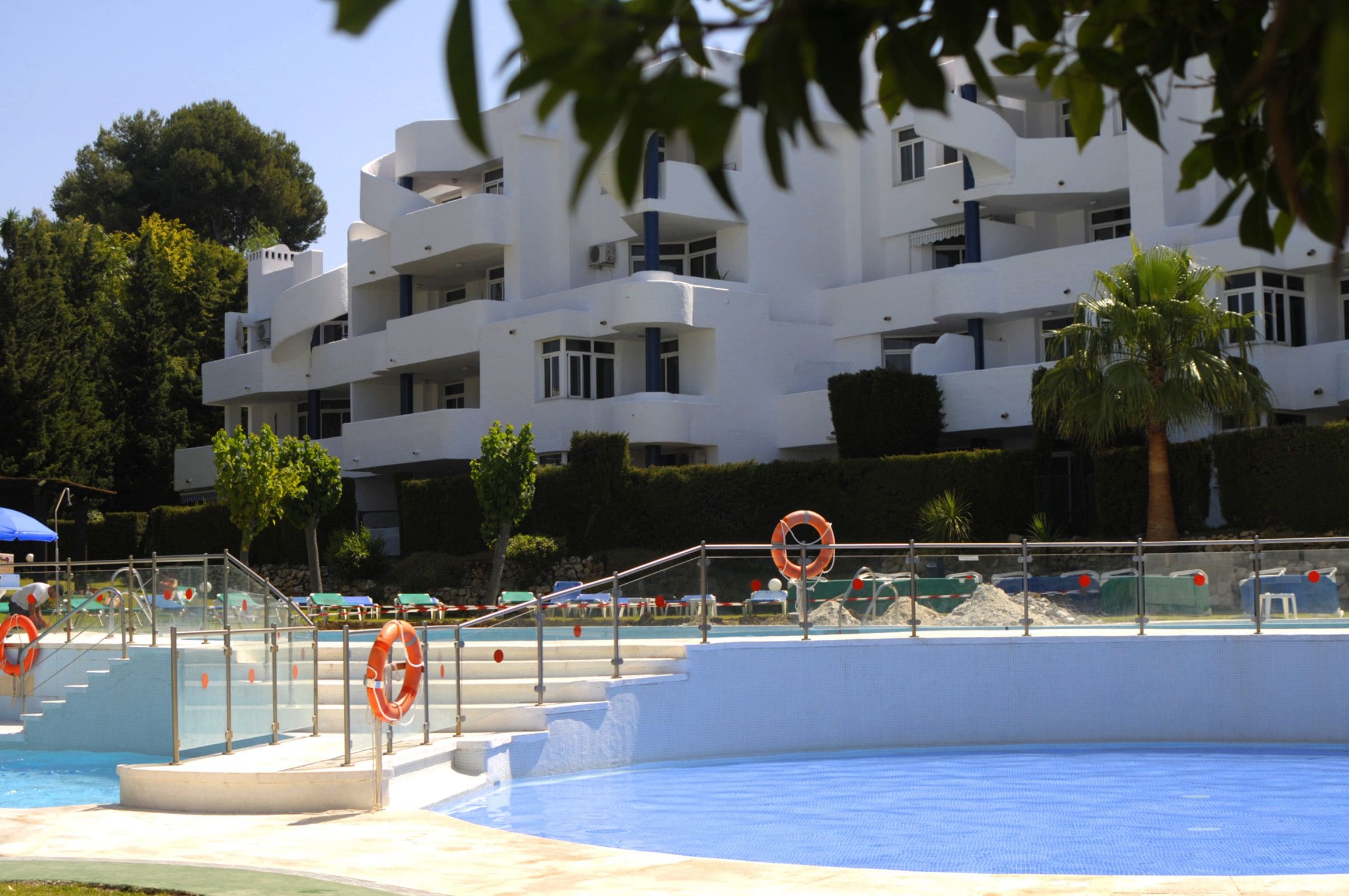 A swimming pool area with a white building in the background, steps leading into the pool, and a palm tree.