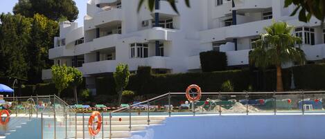 Un área de piscina con un edificio blanco al fondo, escaleras que bajan a la piscina y una palmera.