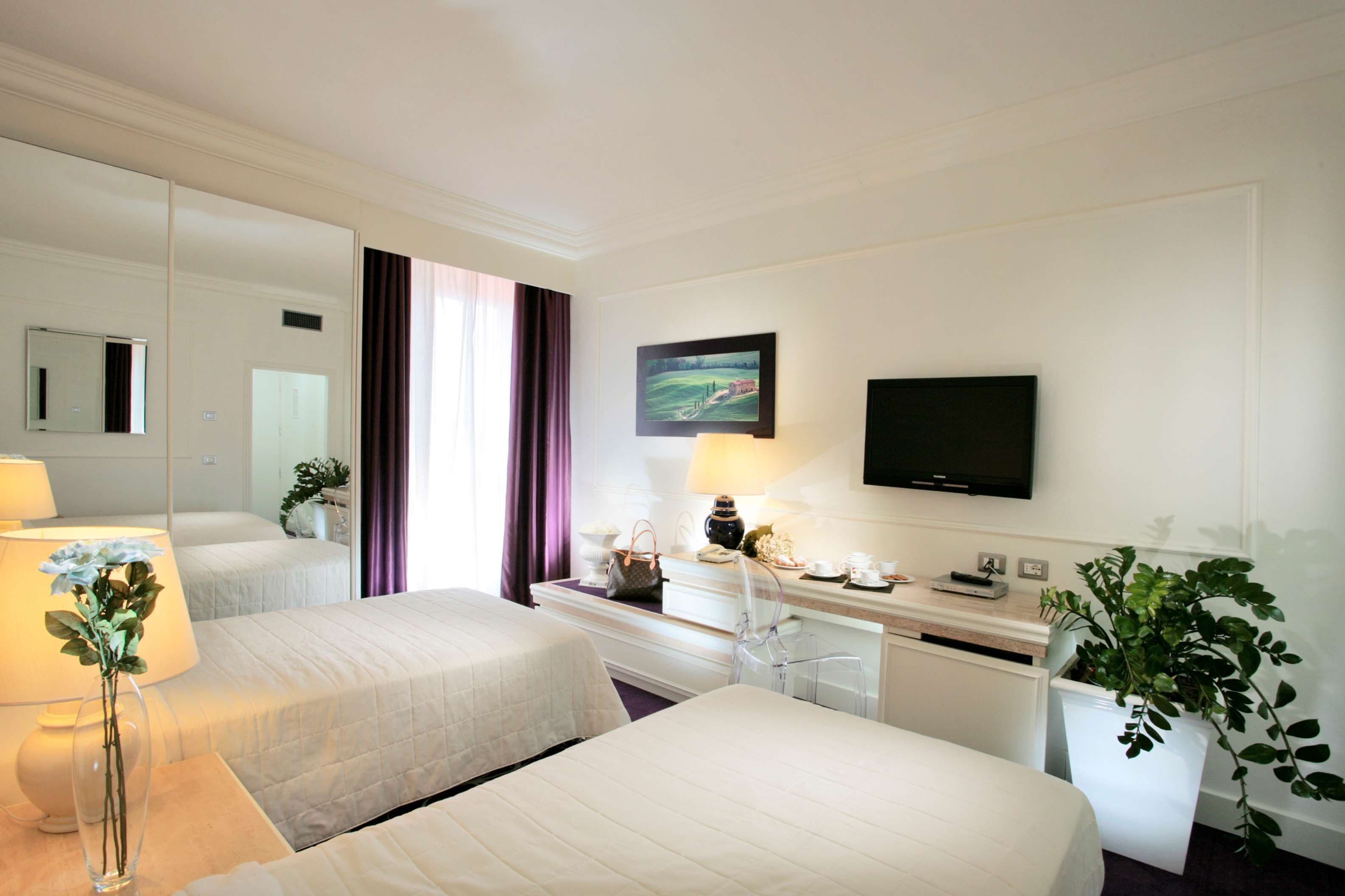 Una camera d'albergo con due letti, una scrivania con televisore, uno specchio e una finestra con tende.