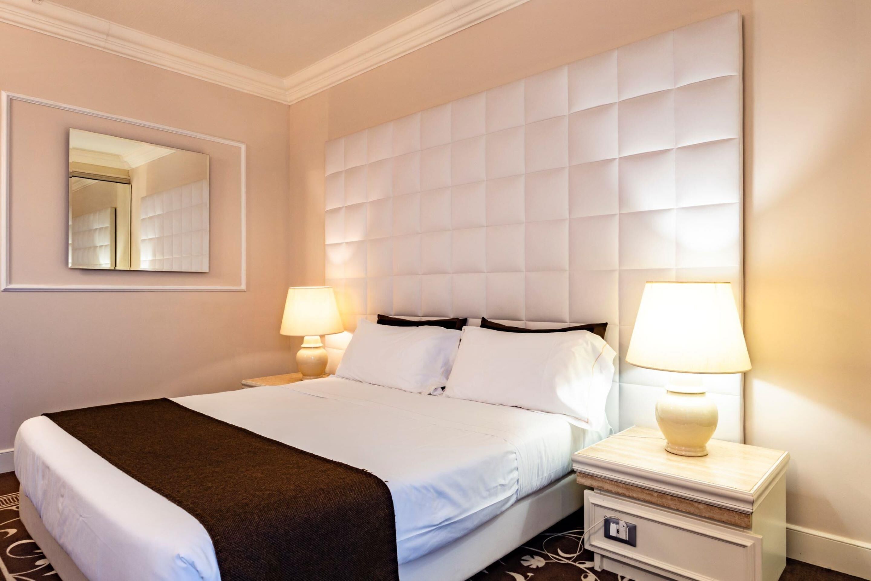 A hotel room with a bed, bedside lamps, a mirror, and a nightstand with a lamp.