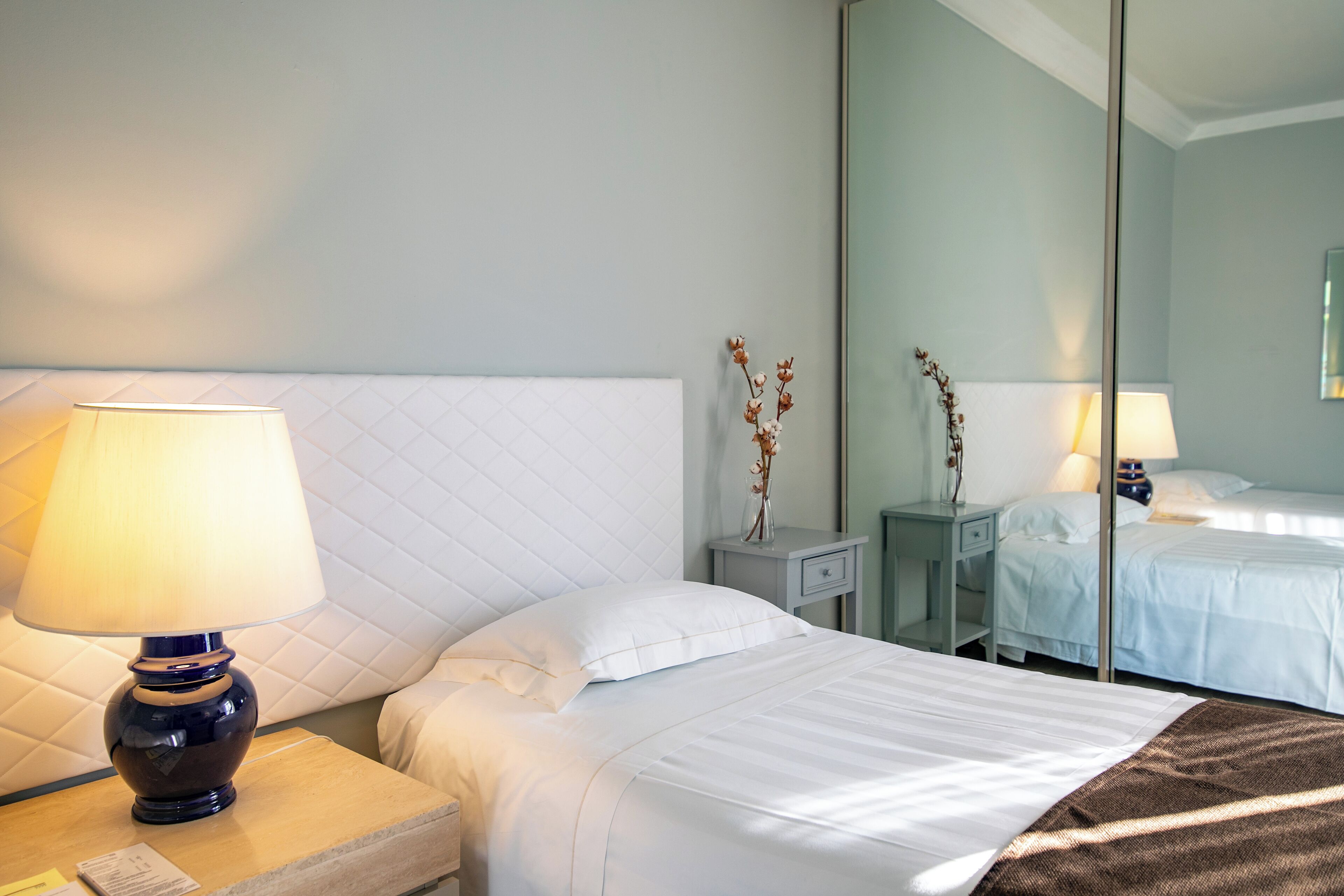 A hotel room with a bed, a nightstand with a lamp, a mirror, and a small table with a vase of dried flowers.
