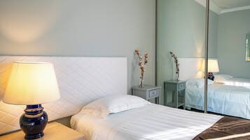 A hotel room with a bed, a nightstand with a lamp, a mirror, and a small table with a vase of dried flowers.