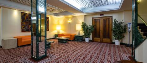 A lobby with patterned carpet, a reception desk, a sofa, a lamp, a potted plant, and a staircase.
