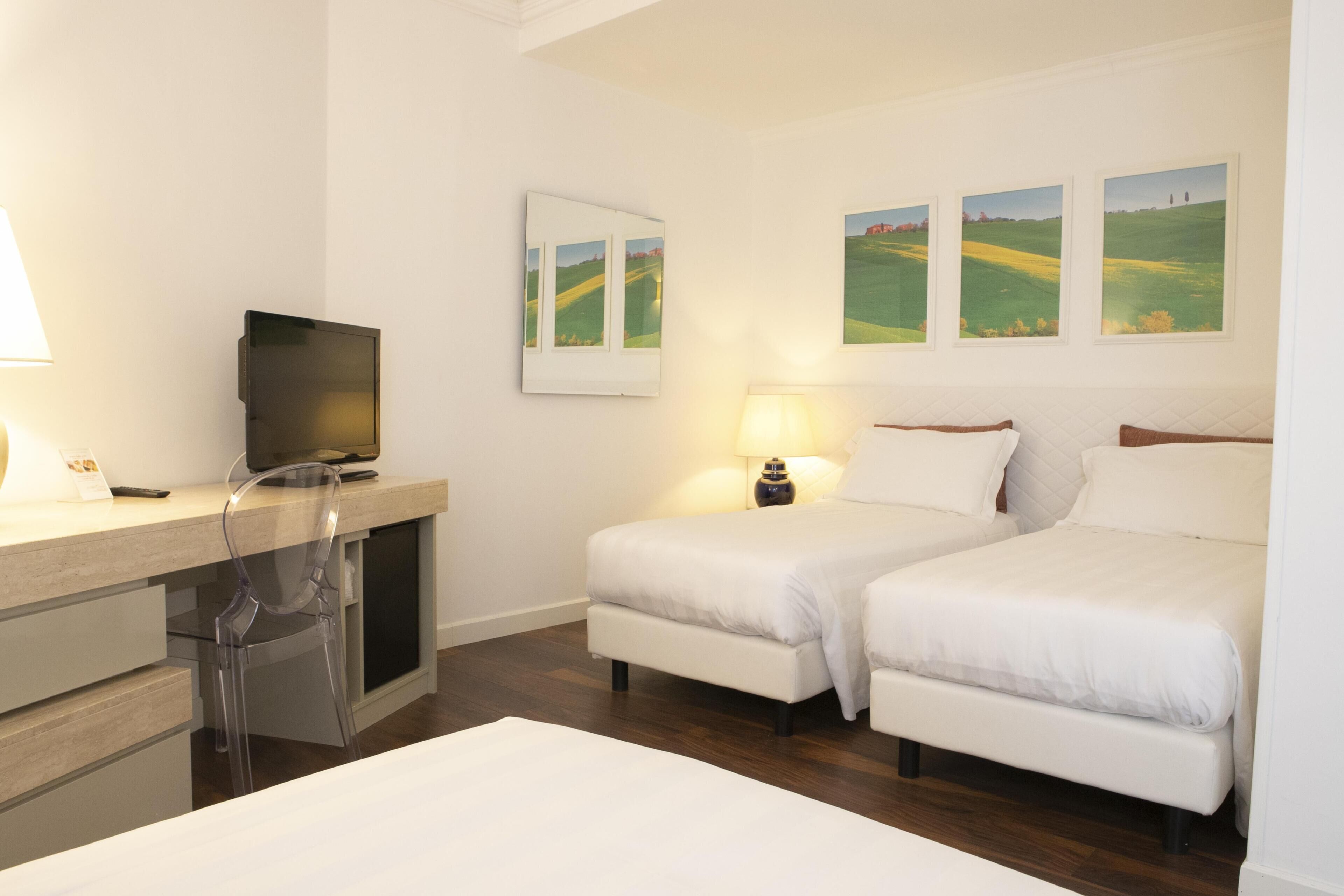 Una habitación de hotel con un escritorio, dos camas, un televisor y tres pinturas de paisajes en la pared.