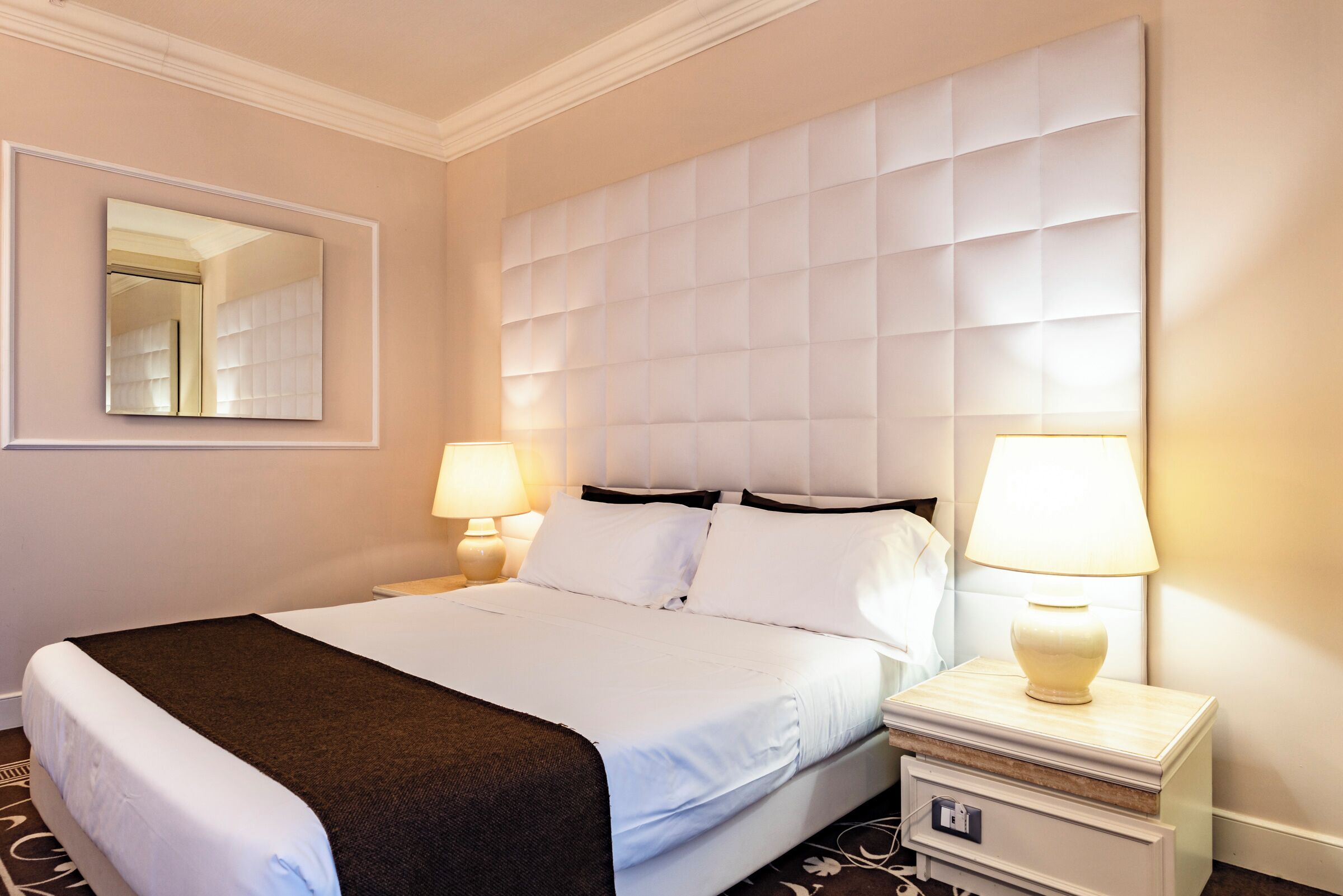 A hotel room with a large bed, two bedside tables with lamps, a mirror, and a padded headboard.