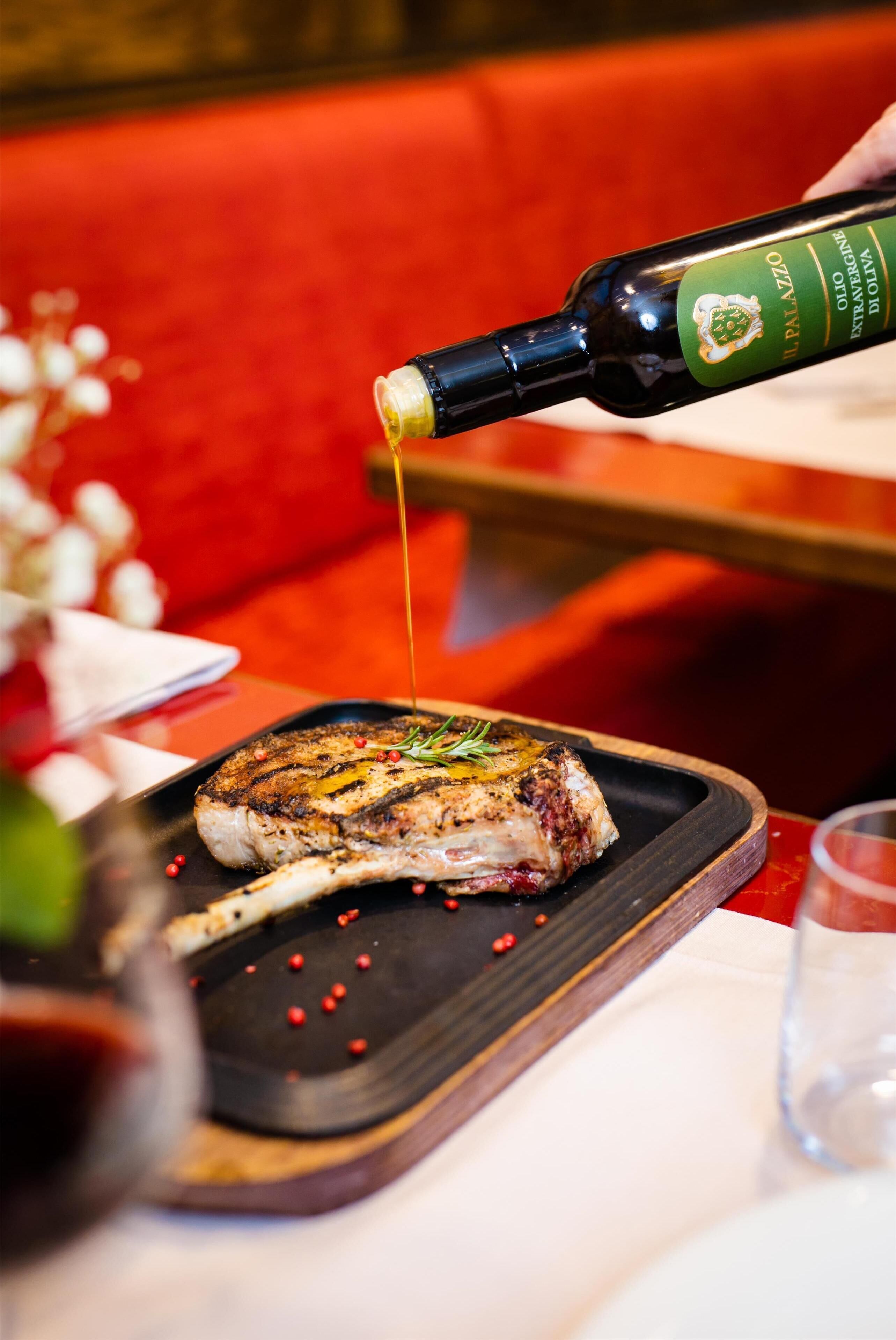 A bottle of olive oil being poured onto a grilled steak on a wooden board.