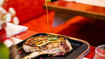 A bottle of olive oil being poured onto a grilled steak on a wooden board.
