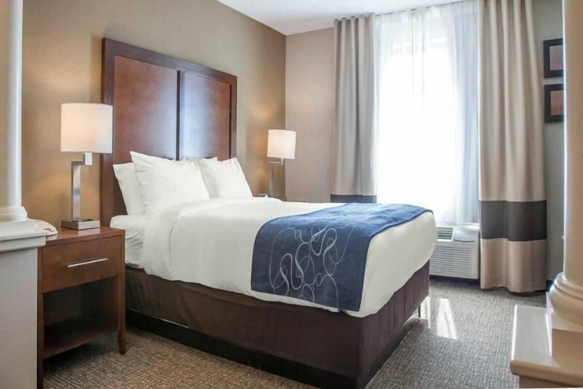 A hotel room with a large bed, two bedside tables, a nightstand, a window with curtains, and a wall-mounted lamp.