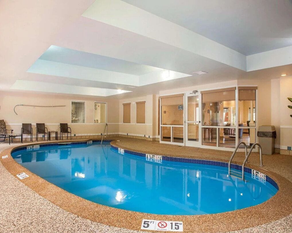 An indoor swimming pool with a 5 ft depth limit, surrounded by a pebble floor and glass doors leading to a room with seating and a counter.
