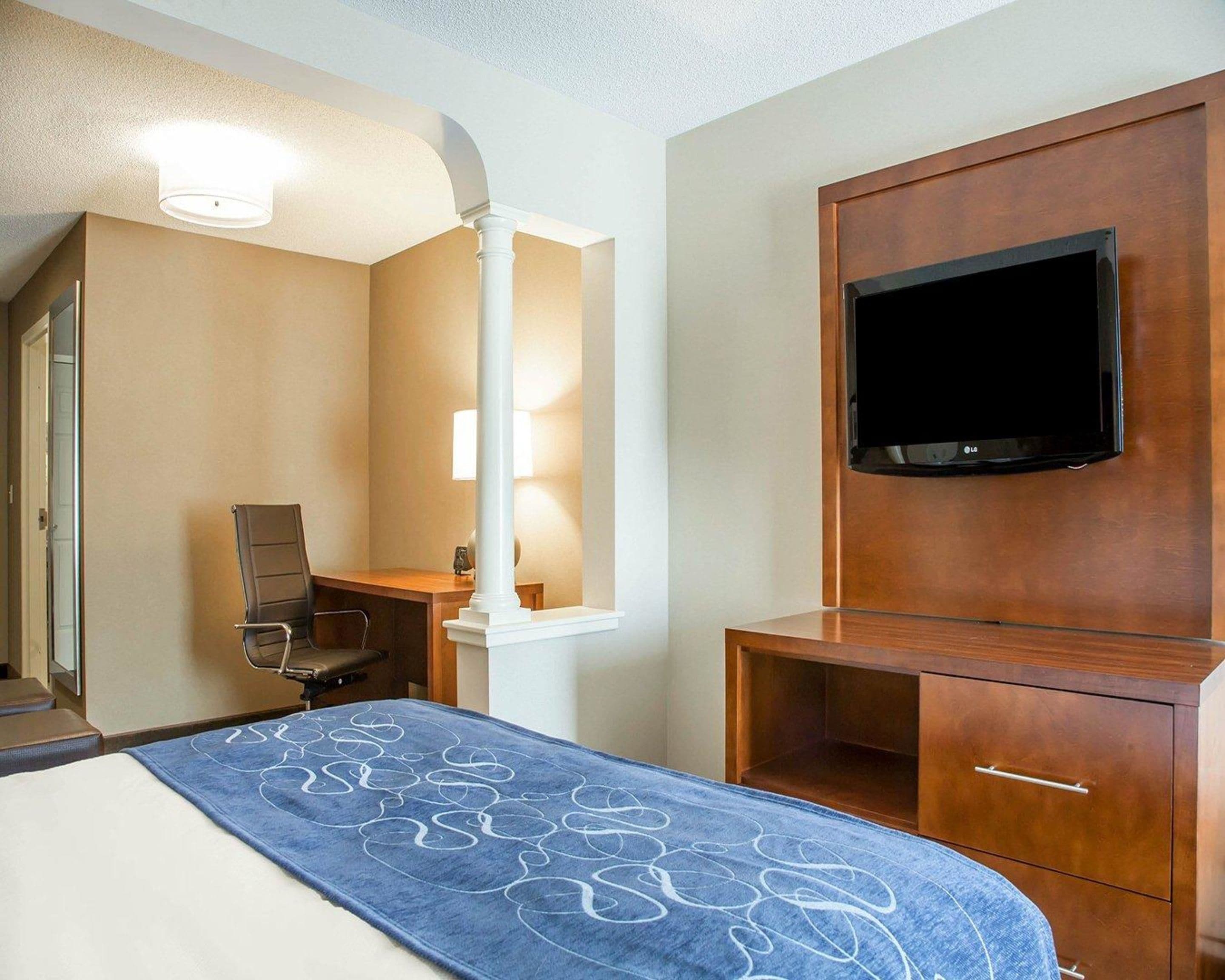 A hotel room with a bed, a desk with a chair, a television mounted on a wooden cabinet, and a mirror with a light above it.