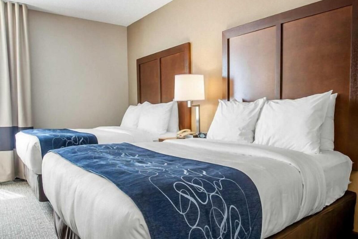 Une chambre d’hôtel avec deux lits, une tête de lit en bois, une table de chevet avec une lampe et une fenêtre avec des rideaux.