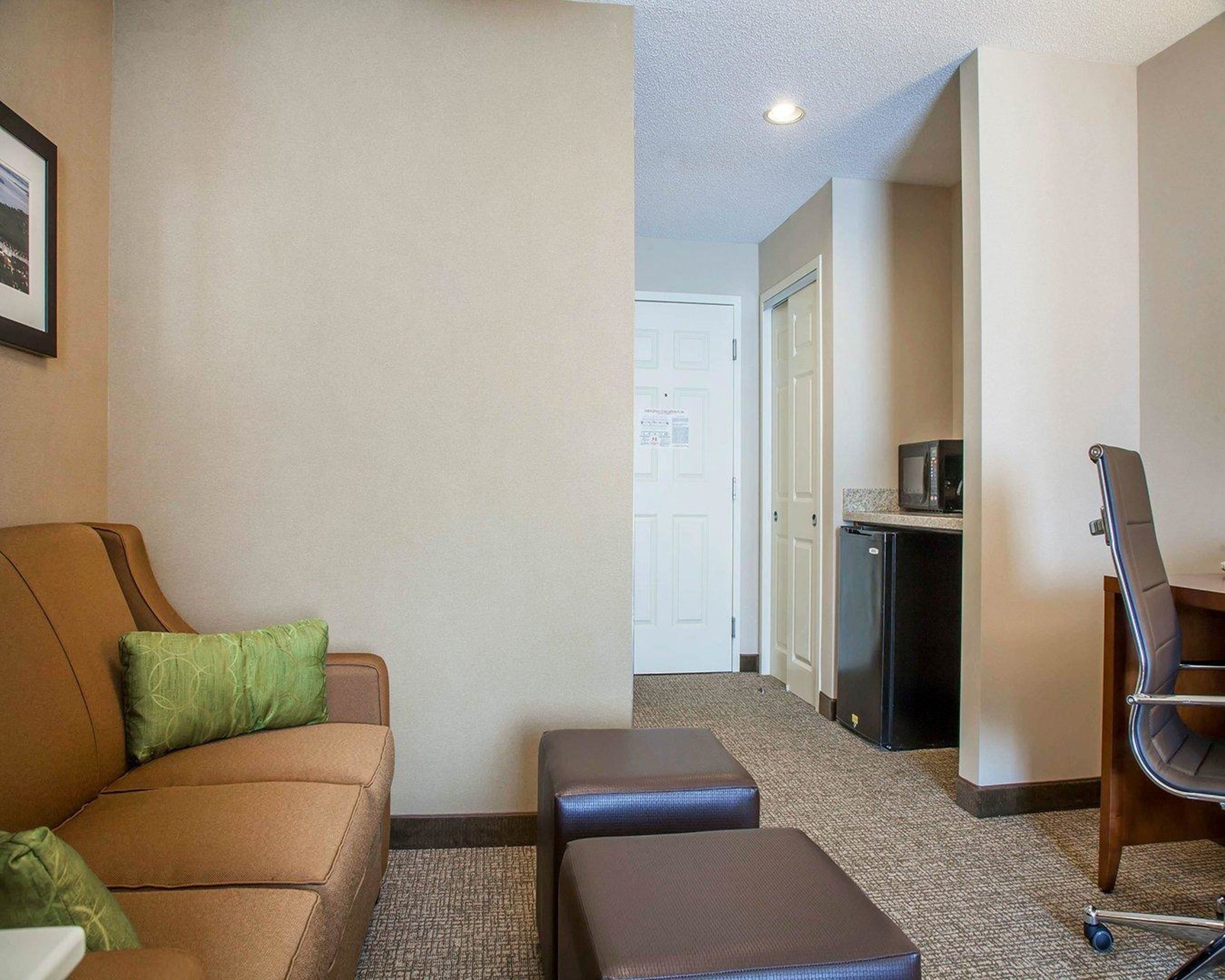 Una habitación de hotel con un sofá marrón, almohada verde y un pequeño reposapiés. Cuenta con una kitchenette equipada con microondas y refrigerador.