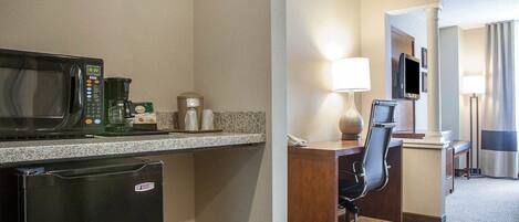 A hotel room with a kitchenette, including a microwave, coffee maker, and refrigerator. There is a desk with a chair and a TV mounted on the wall.