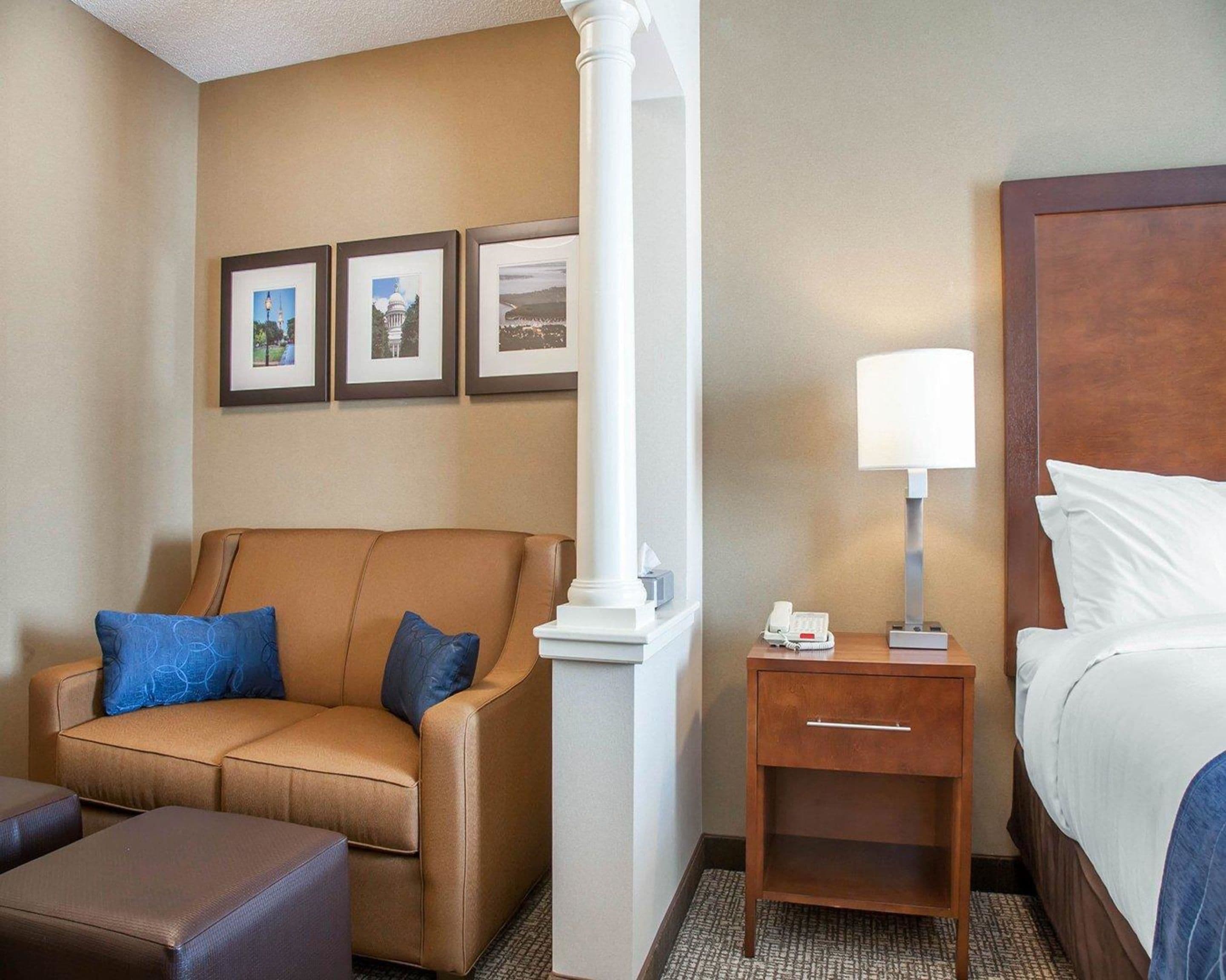 Habitación de hotel con un sofá de cuero marrón, una mesita de noche de madera con una lámpara, una cama con sábanas blancas y tres cuadros en la pared.
